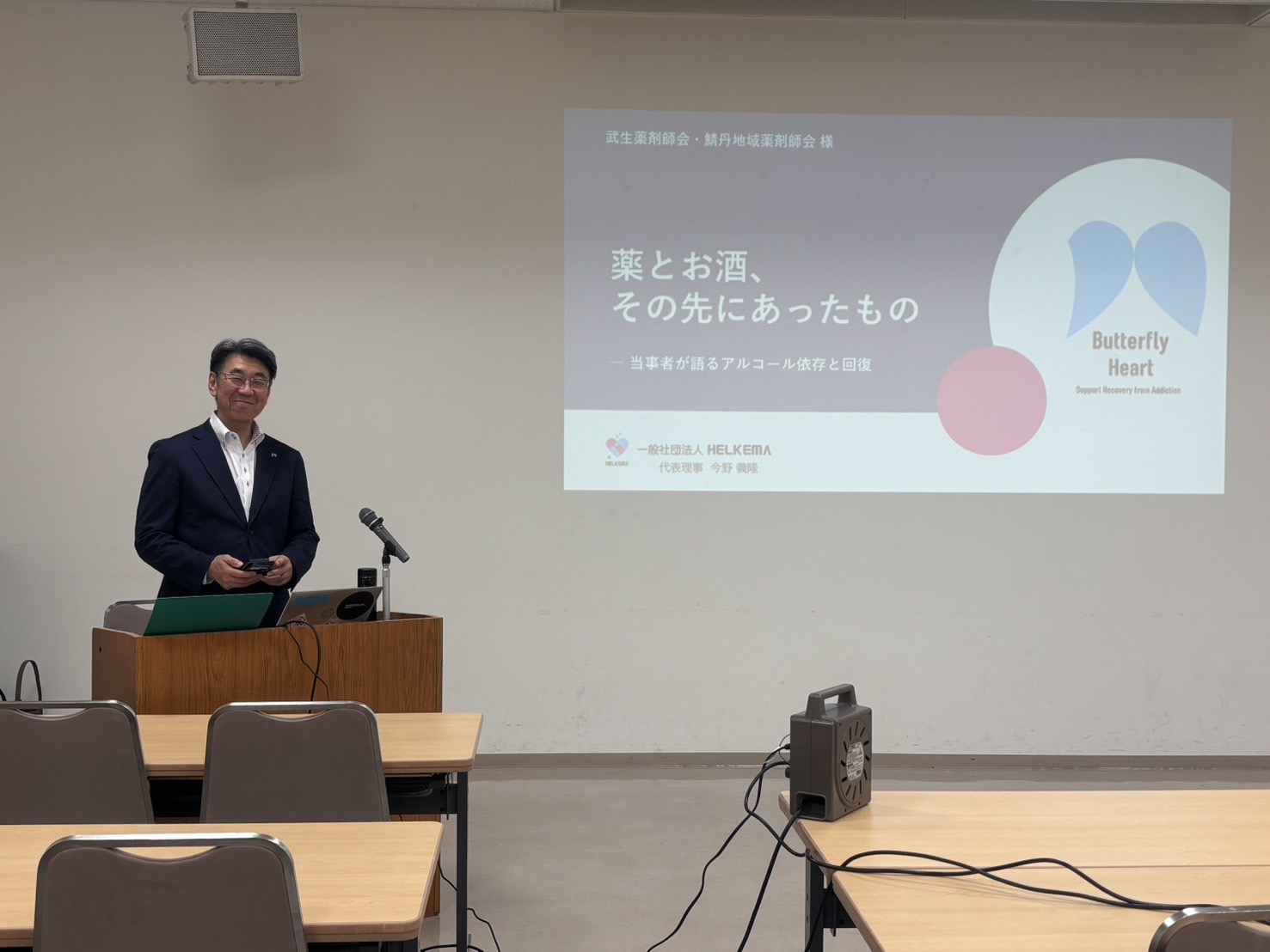 武生薬剤師会・鯖丹地域薬剤師会の講演で、演題を映したスクリーンの横に立つ今野義隆